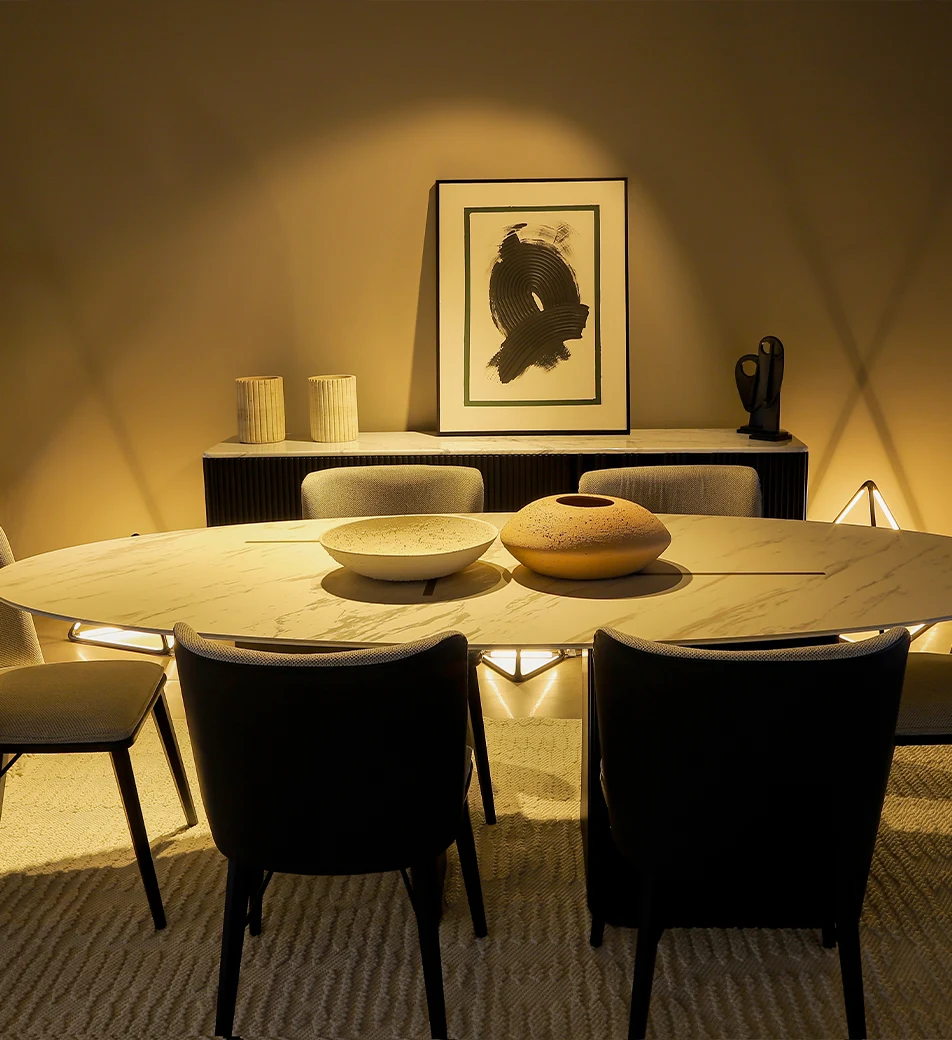A modern dining room with a marble oval table, four chairs, two decorative bowls, abstract art on the wall, and soft yellow lighting creating a cozy atmosphere—perfectly capturing the elegance often showcased in Furniture Photography in Dubai.