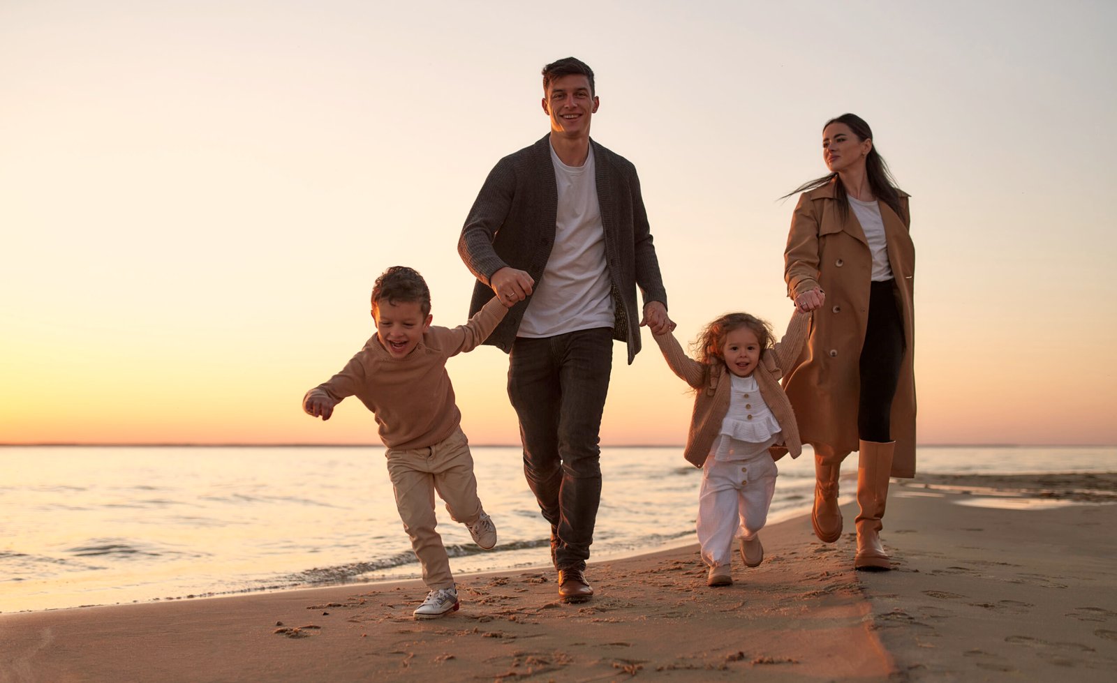 Young family with two children walking on the sandy beach near the sea at sunset having fun and playing.