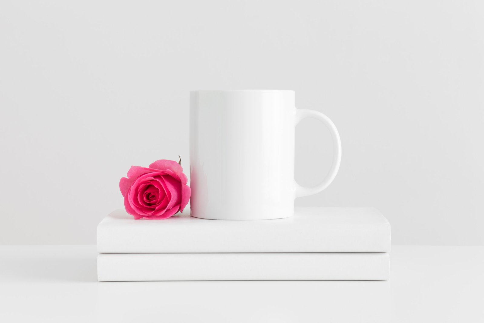 Mug mockup with books and a pink rose on a white table.